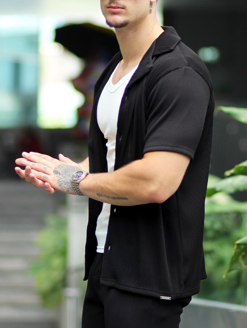 Man wearing a  Balinaro Bailey black  shirt over a white undershirt with tattoos on his arms, standing outdoors.