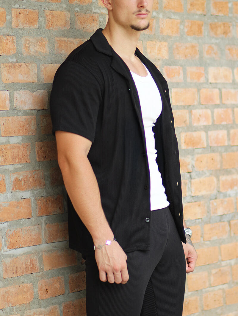 Person wearing a  Balinaro Bailey black  open shirt with black pants against a brick wall.
